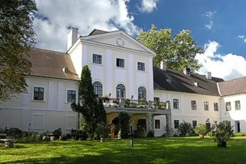 Ein großes weißes Herrenhaus mit einer Uhr, umgeben von saftigem grünem Rasen und Bäumen, unter einem bewölkten blauen Himmel.