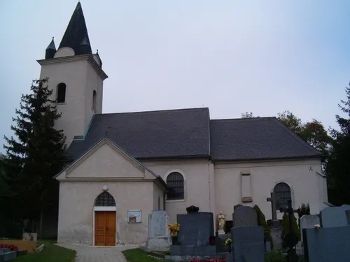 Eine traditionelle Kirche mit einem Turm und einem Schieferdach, gelegen auf einem Friedhof mit Grabsteinen und einer Statue.