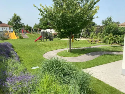 Ein Spielplatz mit Rutsche und Klettergerüst. Der Rasen ist üppig und grün, mit einem Baum in der Mitte. Es gibt Wege und einen Garten mit Blumen.