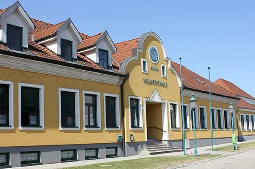 Ein gelbes Gebäude mit einem roten Dach und einer Uhr auf der Vorderseite hat mehrere Fenster und eine Tür. Das Gebäude ist von Gras und Straßenlaternen umgeben.
