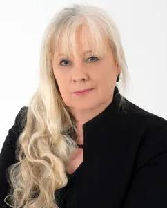 Eine Frau mit langen blonden Haaren, die eine schwarze Jacke trägt, schaut mit ernster Miene in die Kamera.