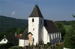Eine weiße Kirche mit schwarzem Turm steht in einer ländlichen Gegend. Sie ist von Bäumen und einem Friedhof umgeben. Ein Berg ist im Hintergrund sichtbar.