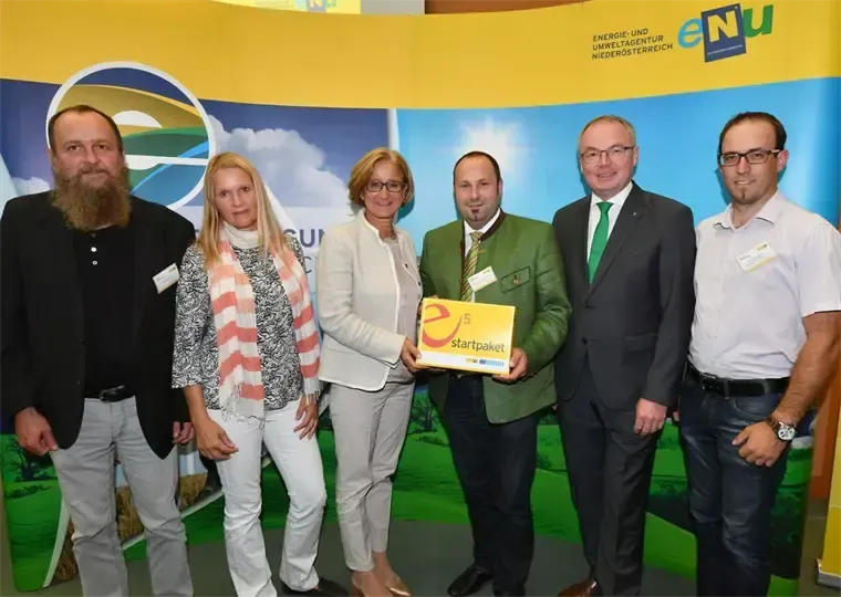 Fünf Erwachsene, drei Männer und zwei Frauen, stehen zusammen. Ein Mann hält eine gelbe Box mit der Aufschrift 'Startpaket'. Sie stehen vor einem Banner mit einem Logo und Text.