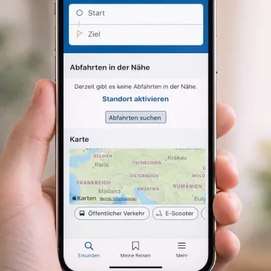 Ein Smartphone-Bildschirm zeigt eine Karte mit mehreren europäischen Ländern. Unter der Karte befinden sich Optionen für 'Öffentlicher Verkehr' und 'E-Scooter'. Der Bildschirm zeigt auch Optionen für 'Entdecken', 'Meine Reisen' und 'Mehr'. Über der Karte ist der Text 'Keine Fahrten in der Nähe' zu sehen.