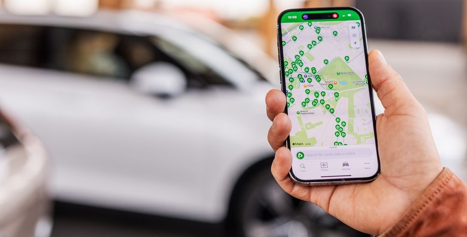 Eine Person hält ein Smartphone mit einer offenen Karten-App, die verschiedene Symbole und eine Suchleiste zeigt. Das Telefon befindet sich vor einem weißen Auto.
