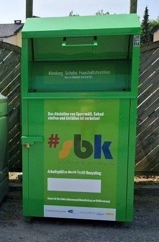 Ein grüner Container für Textilrecycling steht draußen. Er hat einen Schloss und die Wörter #bk darauf. Dahinter ist ein Holzzaun.