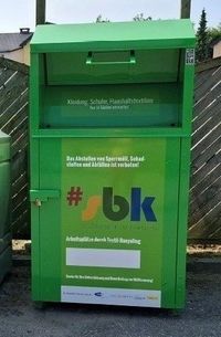 Ein grüner Container für Textilrecycling steht draußen. Er hat einen Schloss und die Wörter #bk darauf. Dahinter ist ein Holzzaun.