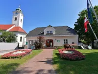 Ein weißes Gebäude mit grauem Dach, Tür und Fenster. Es hat einen Weg mit rosa Blumen und einen Rasen. Links steht ein Uhrturm und rechts eine Fahne.