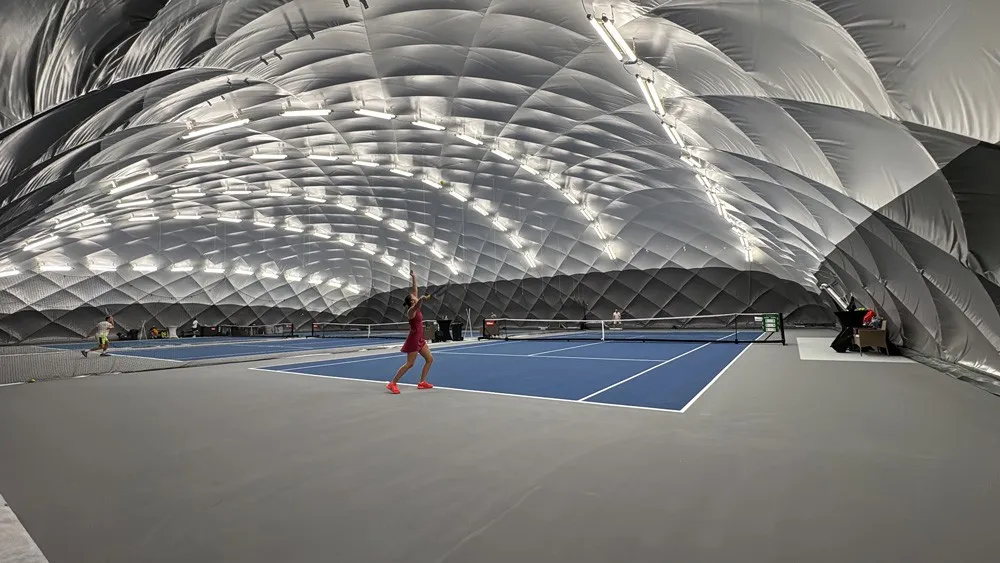 Eine Tennisspielerin befindet sich auf einem Tennisplatz mit blauer Oberfläche, bekleidet mit einem roten Outfit. Es gibt mehrere Tennisplätze und ein Netz ist zwischen ihnen platziert.