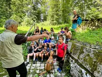 Eine Gruppe von Kindern und Erwachsenen in einem Wald, ein Mann zeigt auf etwas, während ein Hund an der Leine ist.