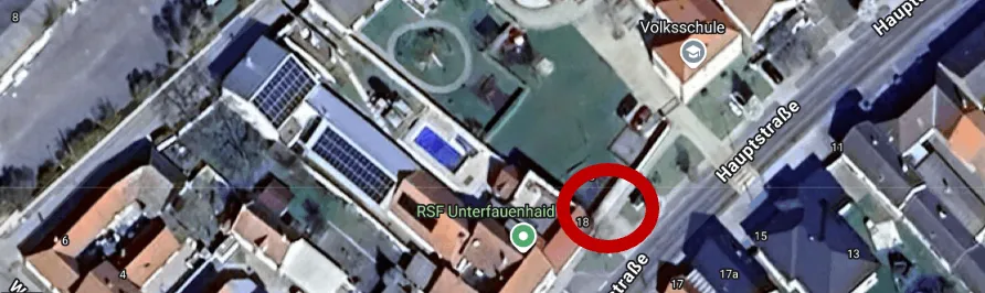 Luftaufnahme eines Wohngebiets mit einem roten Kreis, der ein Gebäude hervorhebt. Der Straßenname 'Hauptstraße' ist sichtbar. Solarmodule sind auf den Dächern installiert.