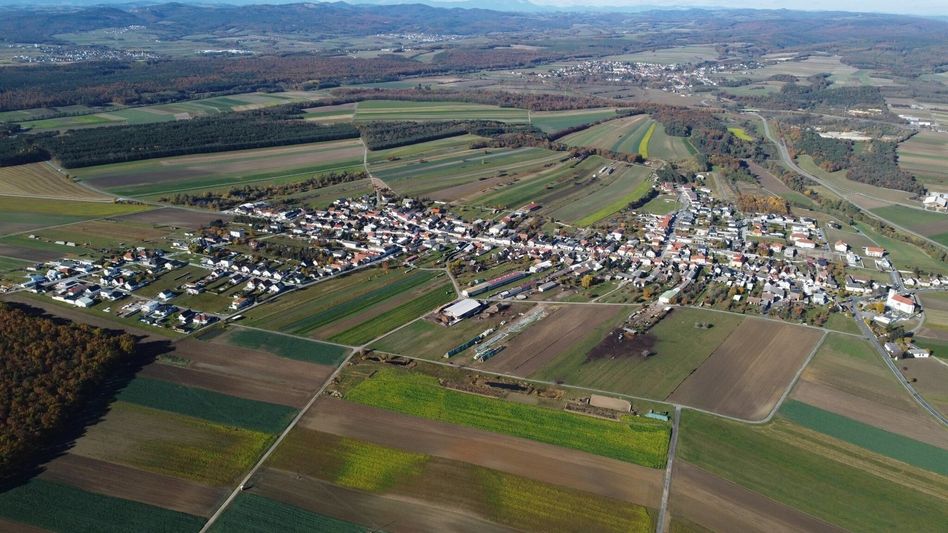 Luftaufnahme einer kleinen Stadt, umgeben von grünen Feldern und Bäumen. Mehrere Häuser und Gebäude sind sichtbar. Die Landschaft ist flach mit entfernten Bergen.
