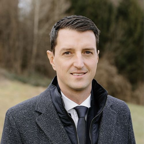 Ein Mann in einem grauen Mantel und einer Krawatte lächelt vor einem Feld mit Bäumen.