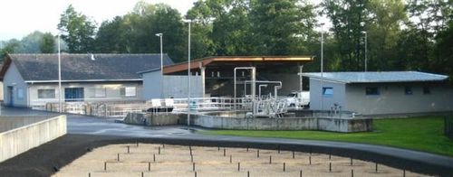 Eine Industrieanlage mit einem großen offenen Bereich davor, umgeben von einer Betonbarriere, und einem Gebäude mit einem geparkten Auto davor.