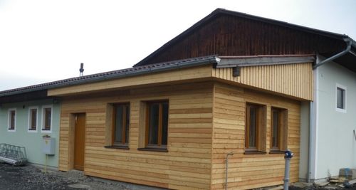 Ein Holzhaus mit brauner Außenseite, mit geneigtem Dach, drei Fenstern, einer Tür und einem Feuerhydranten an der Seite.