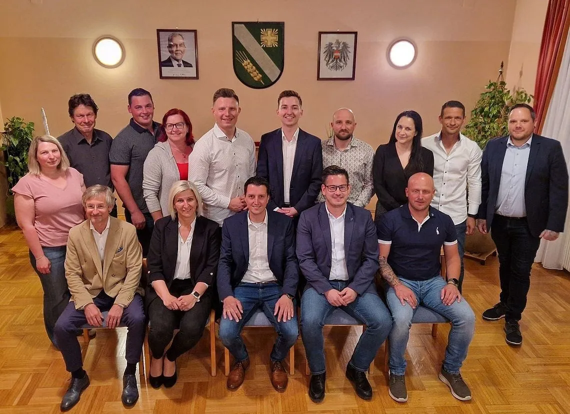 Eine Gruppe Erwachsener posiert für ein Foto im Innenbereich, in formeller und lässiger Kleidung. Dahinter schmücken zwei gerahmte Bilder und ein Schild die Wand. Die Umgebung deutet auf ein professionelles Treffen hin.
