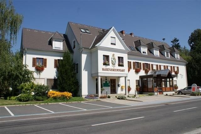Ein großes weißes Gebäude mit braunem Dach, mehreren Fenstern und Pflanzen mit Blumen davor. Es hat ein Schild, das MARKTGENEINPANT steht.
