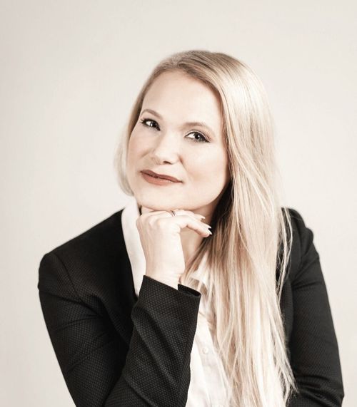 Eine Frau mit langen blonden Haaren trägt einen schwarzen Blazer und ein weißes Hemd. Sie hat einen Ring am Finger und lächelt leicht.