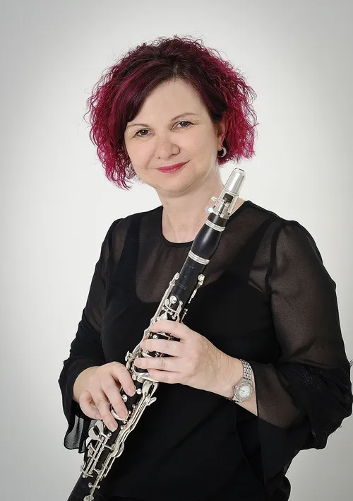 Eine Frau mit roten Haaren lächelt und hält ein Klarinette. Sie trägt ein schwarzes Oberteil und eine Armbanduhr. Der Hintergrund ist weiß.