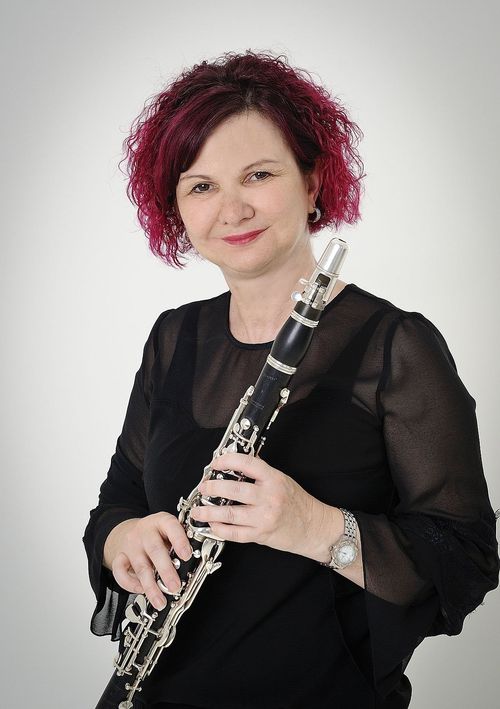 Eine Frau mit roten Haaren lächelt und hält ein Klarinette. Sie trägt ein schwarzes Oberteil und eine Armbanduhr. Der Hintergrund ist weiß.