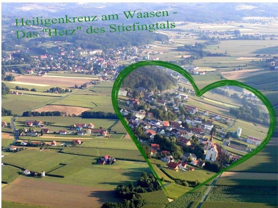 Ein Luftbild eines kleinen Dorfes namens Waasen, mit einem grünen Herz, das das Zentrum hervorhebt. Das Dorf ist von üppigen grünen Feldern und Bäumen umgeben, mit einer Kirche im Zentrum des Herzens.
