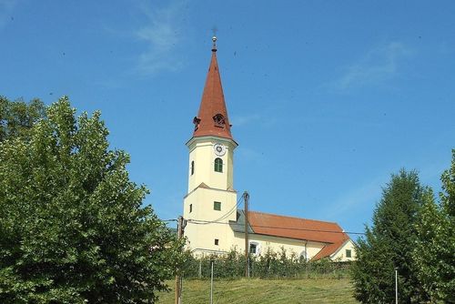 Eine Kirche mit einem Kirchturm und einem roten Dach steht auf einem Hügel. Sie hat eine Uhr auf dem Turm und mehrere Fenster. Die Umgebung ist üppig mit Bäumen und Sträuchern.