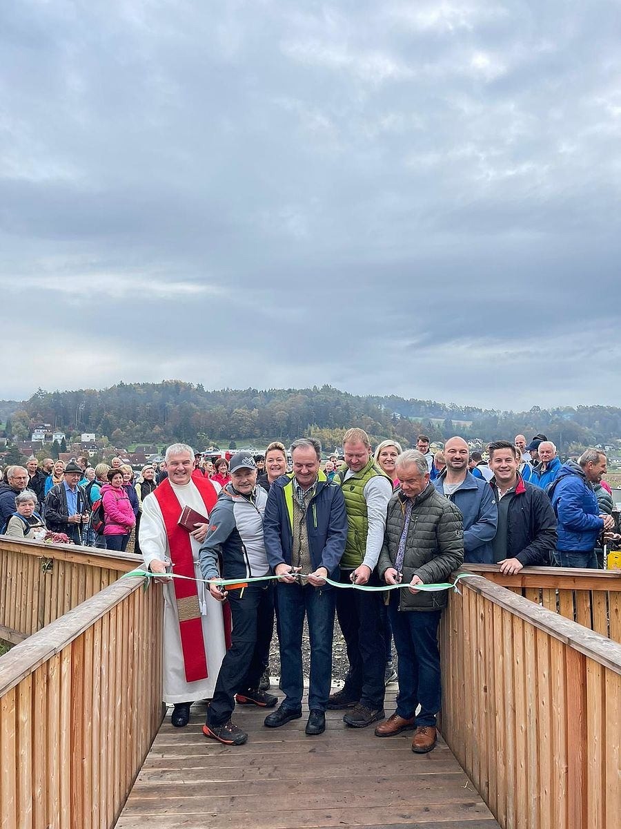 Eine Gruppe von Menschen steht auf einer Holzbrücke. Ein Priester in einem weißen Gewand schneidet ein grünes Band. Hinter ihnen steht eine Menschenmenge und beobachtet. Im Hintergrund ist eine Landschaft mit Bergen und Gebäuden zu sehen.