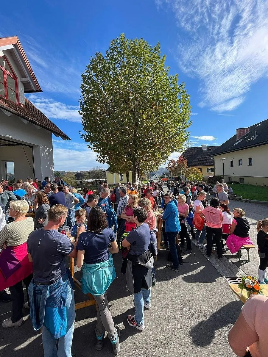 Eine Menschenmenge versammelt sich um Tische unter einem Baum in einem Dorf, einige tragen Schürzen, während ein Kind auf einer Bank steht. Das Gebiet ist von Häusern und Bäumen umgeben.