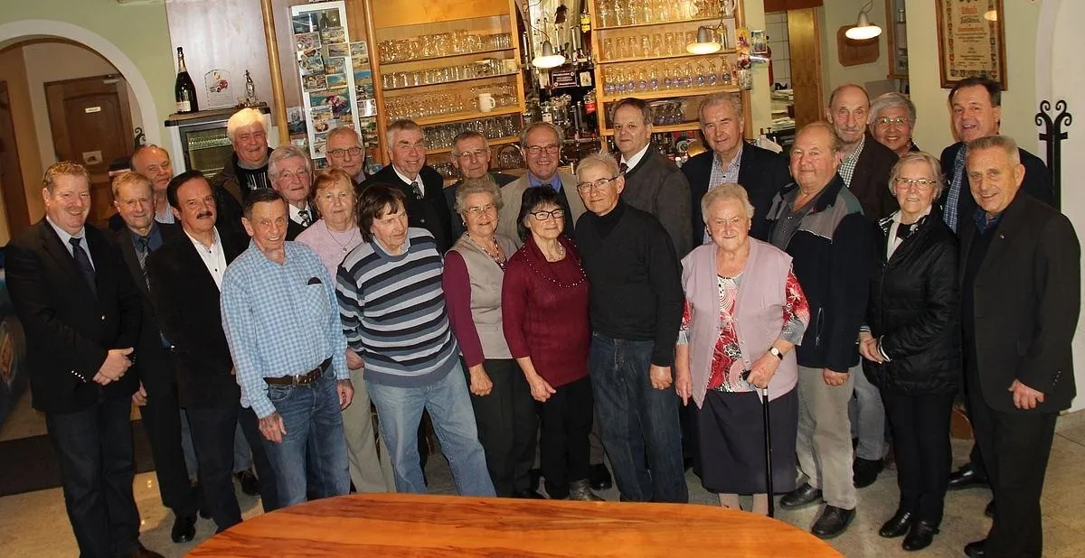 Eine Gruppe älterer Erwachsener versammelt sich in einem Raum, lächelt und posiert für ein Foto. Sie stehen vor einem Holztisch. Der Raum hat Regale, die mit Gläsern und anderen Gegenständen gefüllt sind.