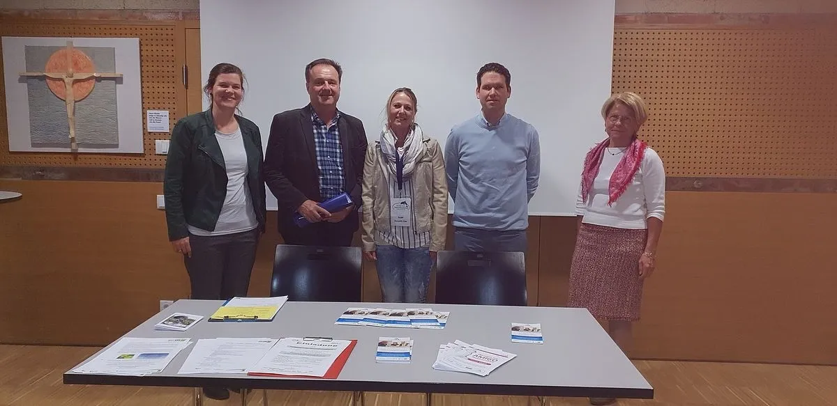 Fünf Personen stehen in einem Raum mit einer weißen Tafel und lächeln für ein Foto. Vier von ihnen stehen hinter einem Tisch mit Papieren, einer hält eine blaue Mappe.