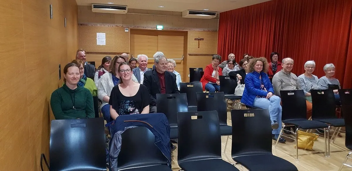 Eine Gruppe von Menschen sitzt in Stühlen in einem Raum, wahrscheinlich in einer Besprechung. Ein Kreuz ist an der Wand hinter den Vorhängen angebracht.