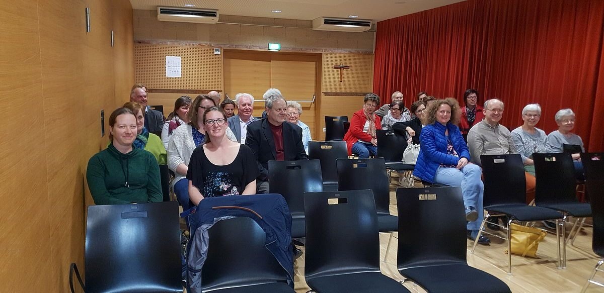 Eine Gruppe von Menschen sitzt in Stühlen in einem Raum, wahrscheinlich in einer Besprechung. Ein Kreuz ist an der Wand hinter den Vorhängen angebracht.