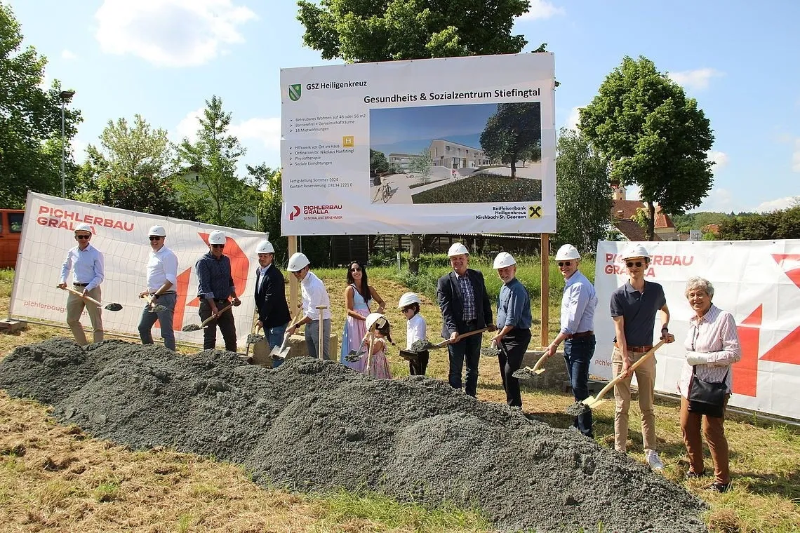 Eine Gruppe von Menschen in weißen Helmen und Sonnenbrillen posiert für ein Foto, während sie Schaufeln auf einer Baustelle halten. Hinter ihnen ist ein Banner mit einem Bild eines Gebäudes und etwas Text.