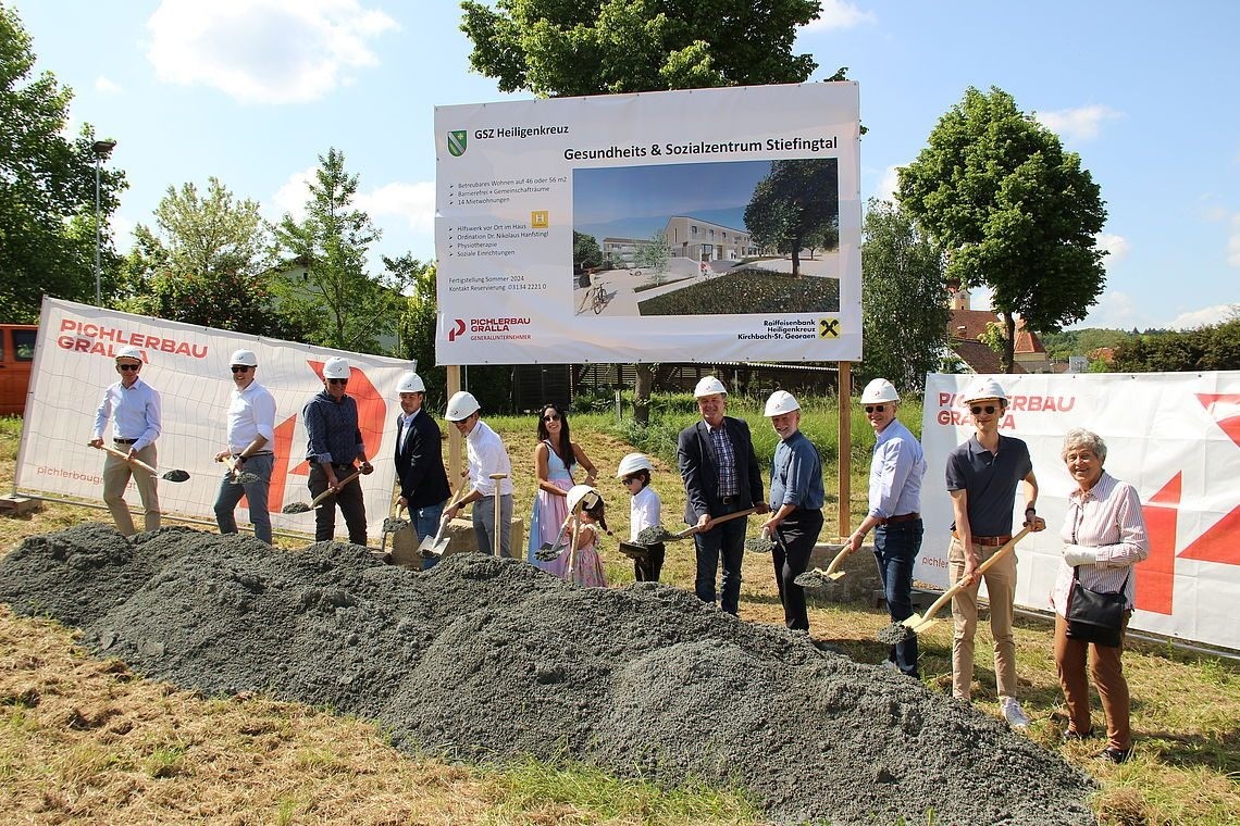 Eine Gruppe von Menschen in weißen Helmen und Sonnenbrillen posiert für ein Foto, während sie Schaufeln auf einer Baustelle halten. Hinter ihnen ist ein Banner mit einem Bild eines Gebäudes und etwas Text.