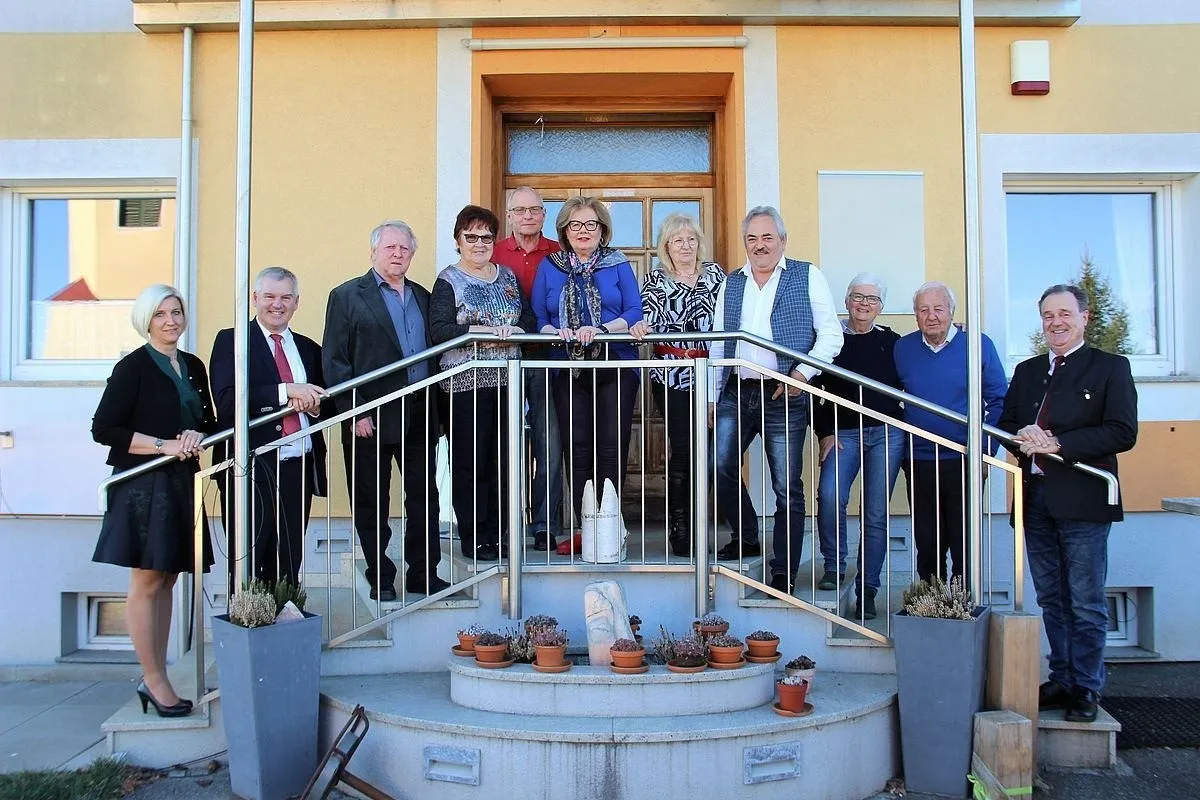 Eine Gruppe Erwachsener steht auf den Stufen vor einem gelben Gebäude. Sie lächeln und schauen nach vorne. Es gibt Topfpflanzen auf den Stufen.