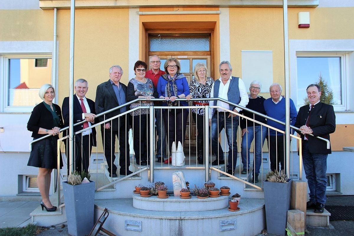 Eine Gruppe Erwachsener steht auf den Stufen vor einem gelben Gebäude. Sie lächeln und schauen nach vorne. Es gibt Topfpflanzen auf den Stufen.