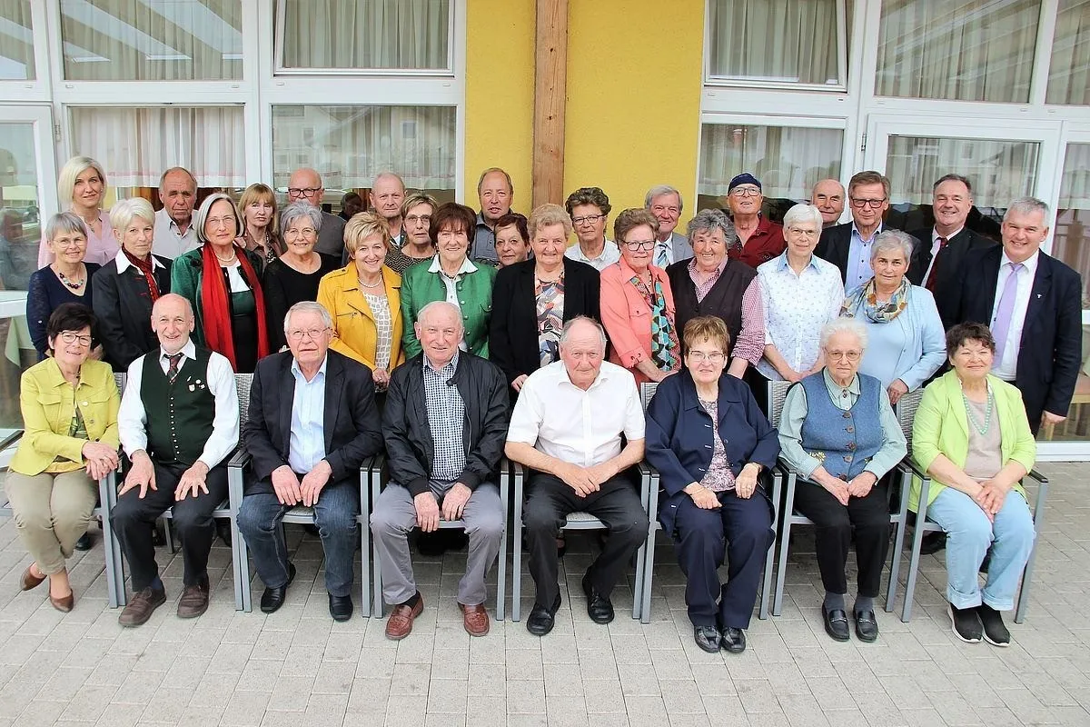 Eine Gruppe älterer Menschen, sowohl Männer als auch Frauen, sitzen und stehen vor einem gelben Gebäude mit weißen Fenstern und Vorhängen. Sie sind in verschiedenen Outfits und Accessoires gekleidet, einige mit Brillen und Hüten.