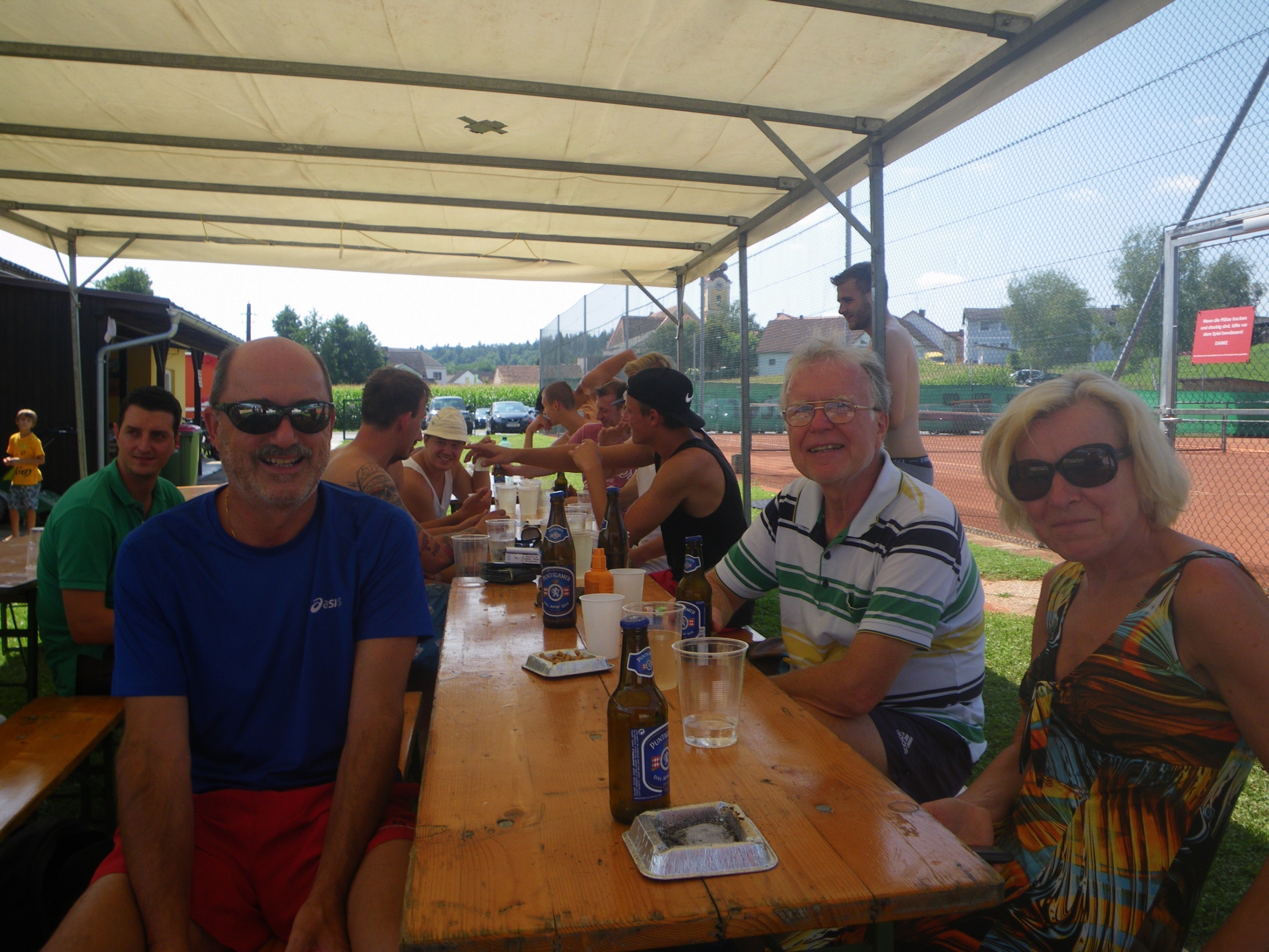Eine Gruppe von Menschen sitzt unter einem Zelt an einem Tisch, einige tragen Sonnenbrillen und Hüte. Flaschen und Gläser stehen auf dem Tisch. Im Hintergrund befinden sich Tennisplätze und Gebäude.