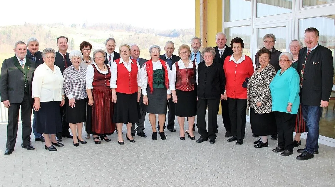 Eine Gruppe von Menschen, darunter Männer und Frauen, steht vor einem Gebäude, wahrscheinlich für ein Gruppenfoto. Sie tragen formelle Kleidung, und einige tragen Brillen. Hinter ihnen ist ein Blick auf die Berge.