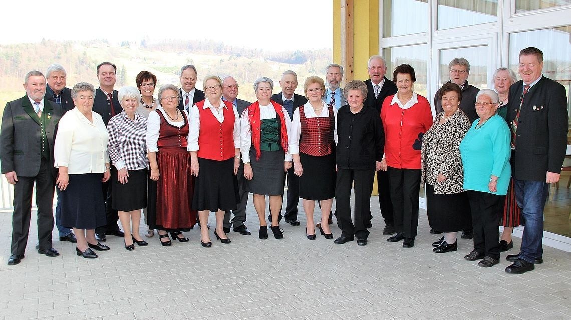Eine Gruppe von Menschen, darunter Männer und Frauen, steht vor einem Gebäude, wahrscheinlich für ein Gruppenfoto. Sie tragen formelle Kleidung, und einige tragen Brillen. Hinter ihnen ist ein Blick auf die Berge.