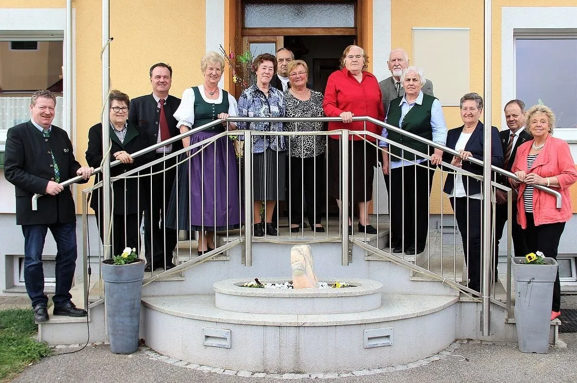 Eine Gruppe von Menschen in verschiedenen Kleidungsstücken, die auf einer Treppe vor einem Gebäude stehen. Links steht ein Topf mit Blumen.