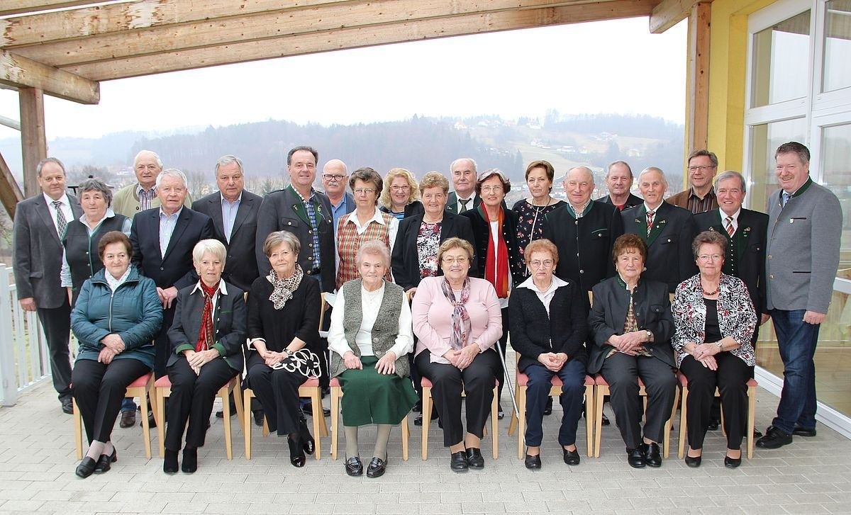 Eine Gruppe älterer Menschen in formeller Kleidung posiert für ein Foto unter einer Holzpergola mit Blick auf Berge und Gebäude im Hintergrund.