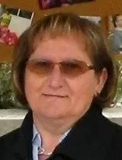 Eine Frau mit kurzen, hellbraunen Haaren, die eine Brille und einen schwarzen Mantel trägt, steht draußen. Hinter ihr ist eine Wand mit Fotos zu sehen.