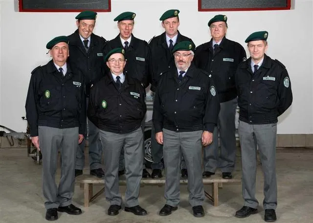 Eine Gruppe uniformierter Personen in schwarzen Jacken und grünen Baretten steht in einer Reihe, lächelt und posiert für ein Foto.