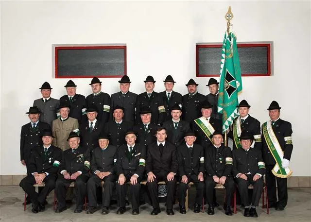Eine Gruppe von Menschen in Uniform posiert für ein Foto vor einer weißen Wand. Sie tragen Hüte, Krawatten und einige haben Medaillen. Hinter ihnen steht eine grüne Flagge und ein grüner Banner.