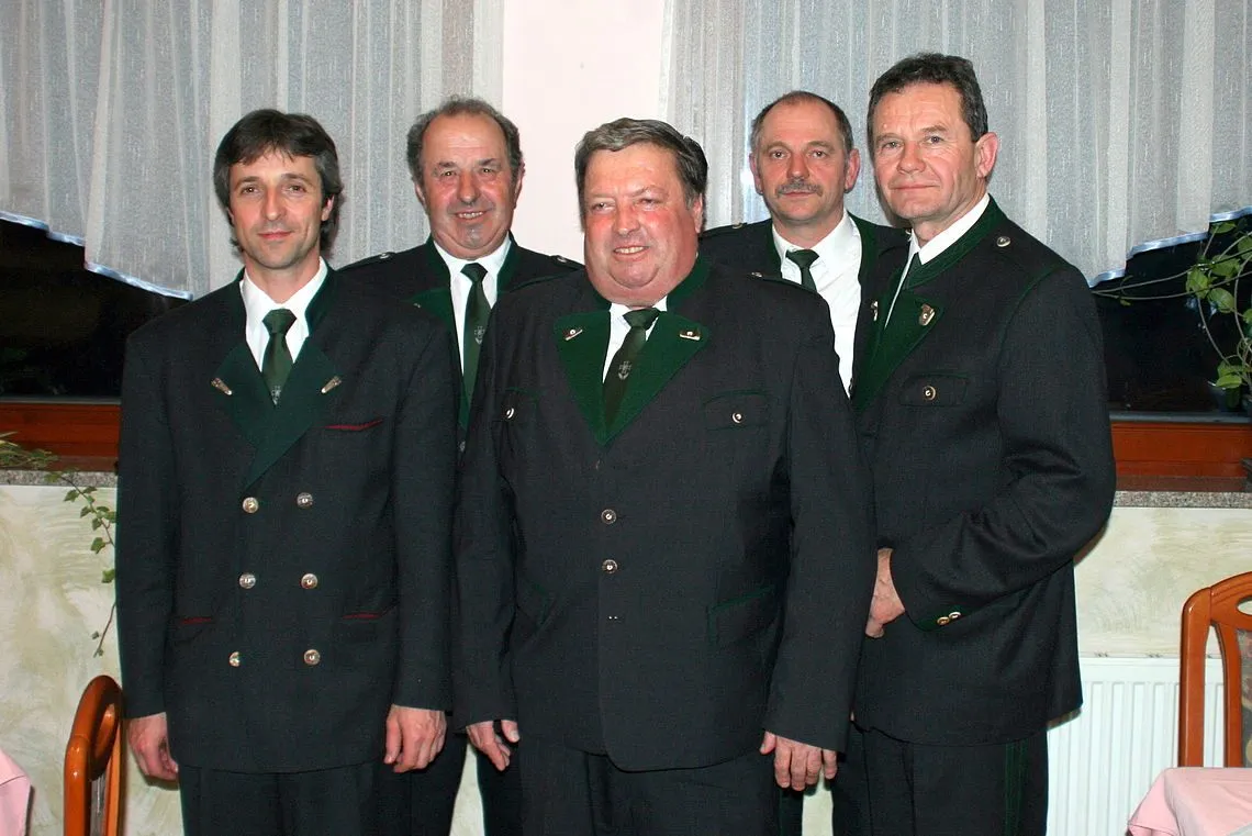 Fünf Männer in übereinstimmenden Uniformen posieren für ein Foto. Sie tragen schwarze Anzüge mit grünen Krawatten und weißen Hemden. Der Mann in der Mitte lächelt und hat leicht geöffneten Mund.