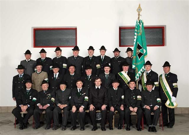Eine Gruppe von Menschen in Uniform posiert für ein Foto vor einer weißen Wand. Sie tragen Hüte, Krawatten und einige haben Medaillen. Hinter ihnen steht eine grüne Flagge und ein grüner Banner.