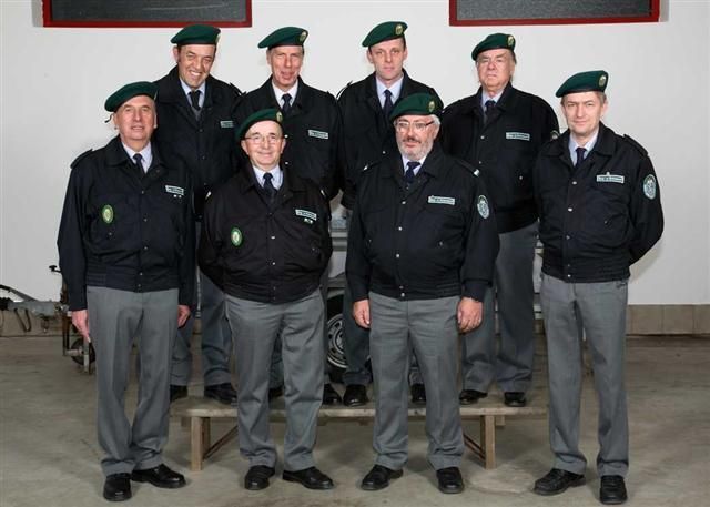 Eine Gruppe uniformierter Personen in schwarzen Jacken und grünen Baretten steht in einer Reihe, lächelt und posiert für ein Foto.