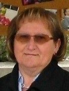 Eine Frau mit kurzen, hellbraunen Haaren, die eine Brille und einen schwarzen Mantel trägt, steht draußen. Hinter ihr ist eine Wand mit Fotos zu sehen.