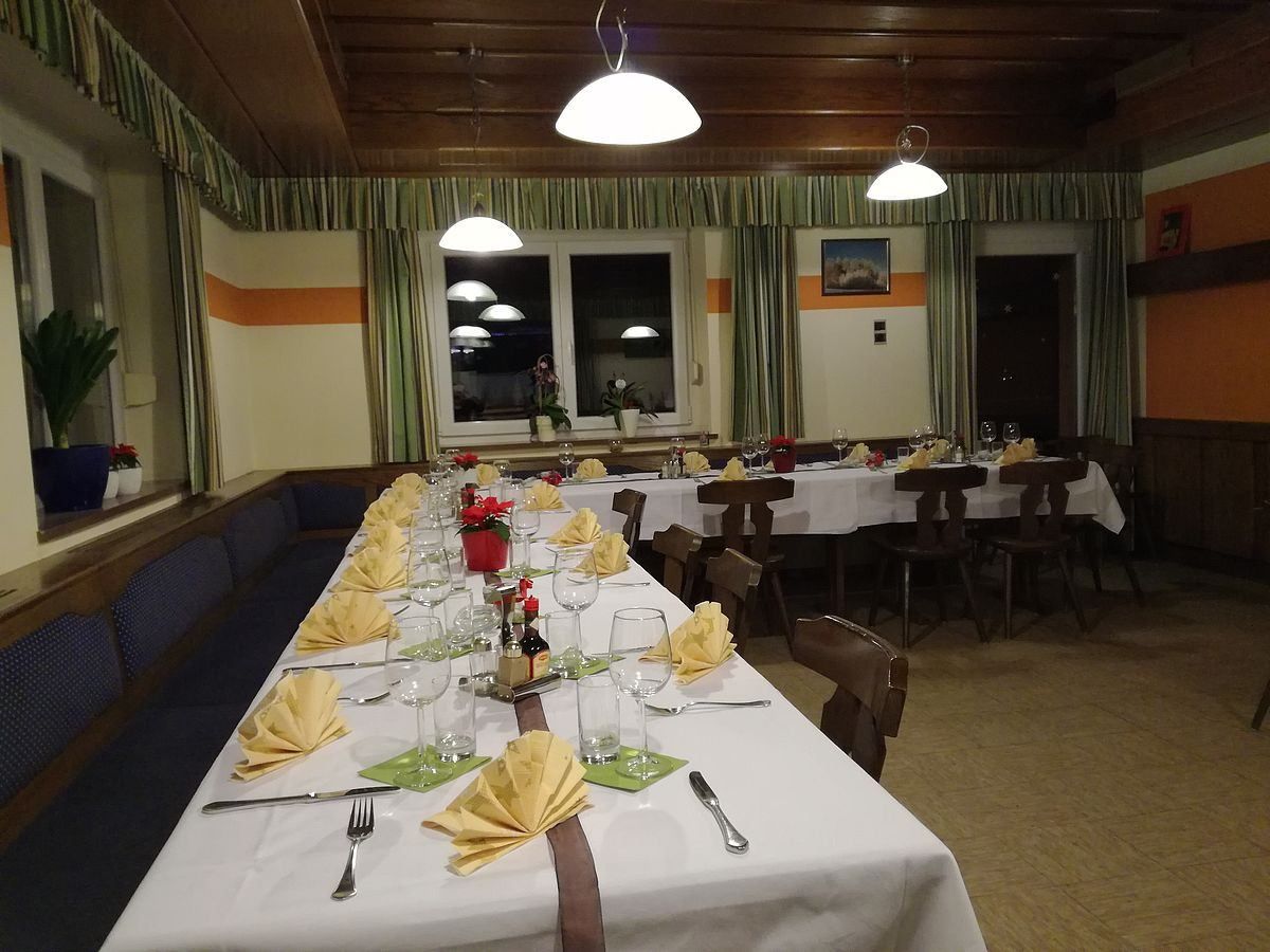 Ein langer Tisch in einem Speisesaal mit ordentlich arrangierten Tischgedecken, einschließlich gefalteter Servietten, Weingläsern und Besteck. Der Raum hat Holzdecken mit hängenden Lampen, einem Bilderrahmen und Fenstern mit Vorhängen.